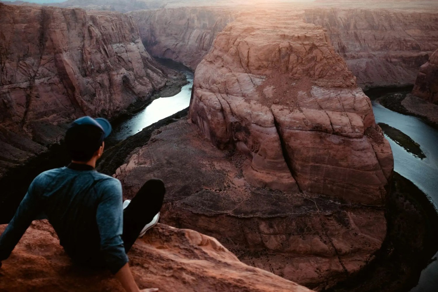 Person in cap sitting on cliff overlooking horseshoe bend river canyon at sunset.