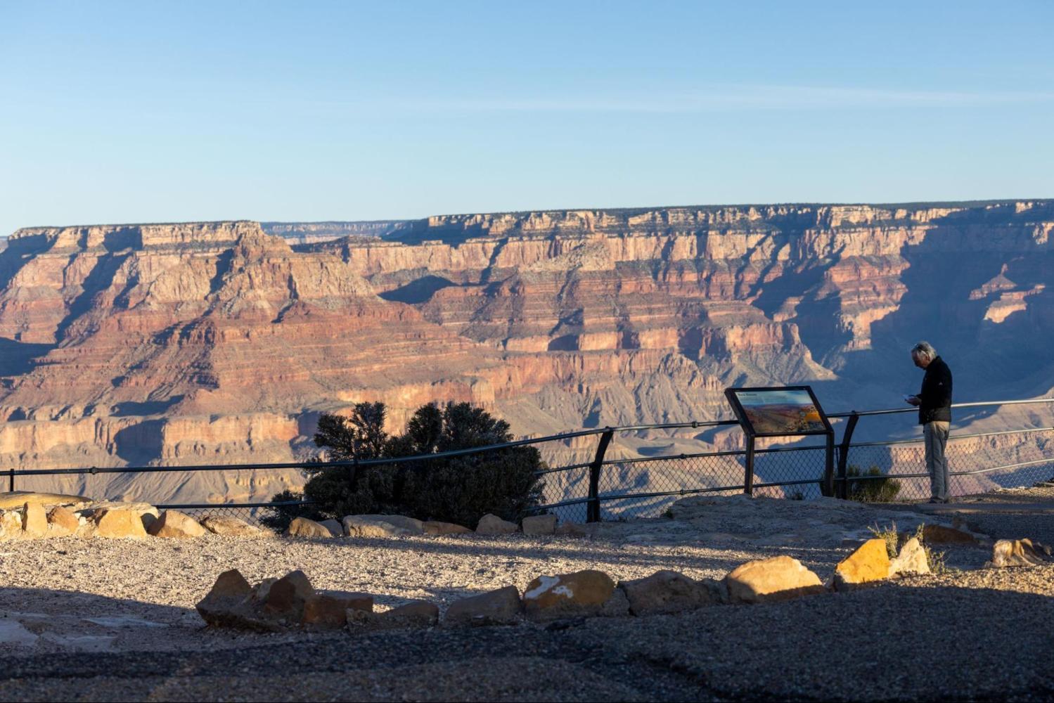 Person reading sign at Grand Canyon viewpoint under clear sky.